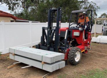 A worker operating a red forklift moving bundled concrete posts at a construction site