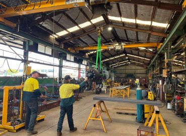 Workers in high-vis clothing using an overhead crane to lift a heavy industrial component inside a workshop