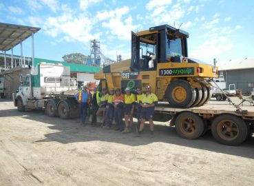 A group of workers standing next to a low-loader carrying a Caterpillar compactor, representing teamwork in machinery logistics