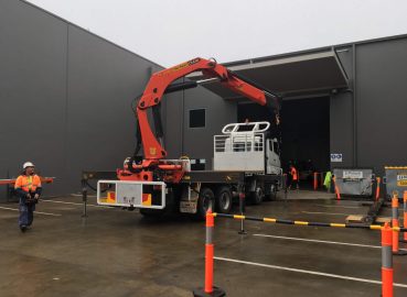 A truck-mounted Palfinger crane lifting materials outside an industrial facility, with workers in high-vis gear ensuring safety