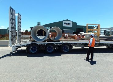 Concrete Pipe Delivery at Humes Facility – Workers securing and inspecting large concrete pipes on a transport truck outside the Humes facility