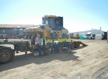 A group of workers in high-vis uniforms standing in front of a low-loader trailer carrying a large Caterpillar compactor, representing heavy machinery logistics