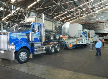 A blue Kenworth truck hauling industrial equipment inside a warehouse, with workers monitoring the process